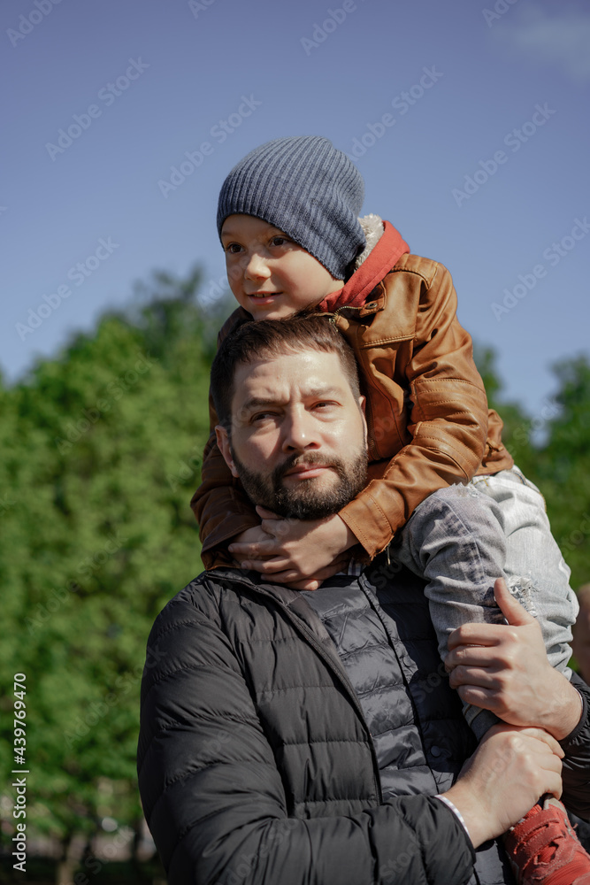 young handsome caucasian bearded man holding on his shoulder 7 year old son. Father and son in park. Image with selective focus