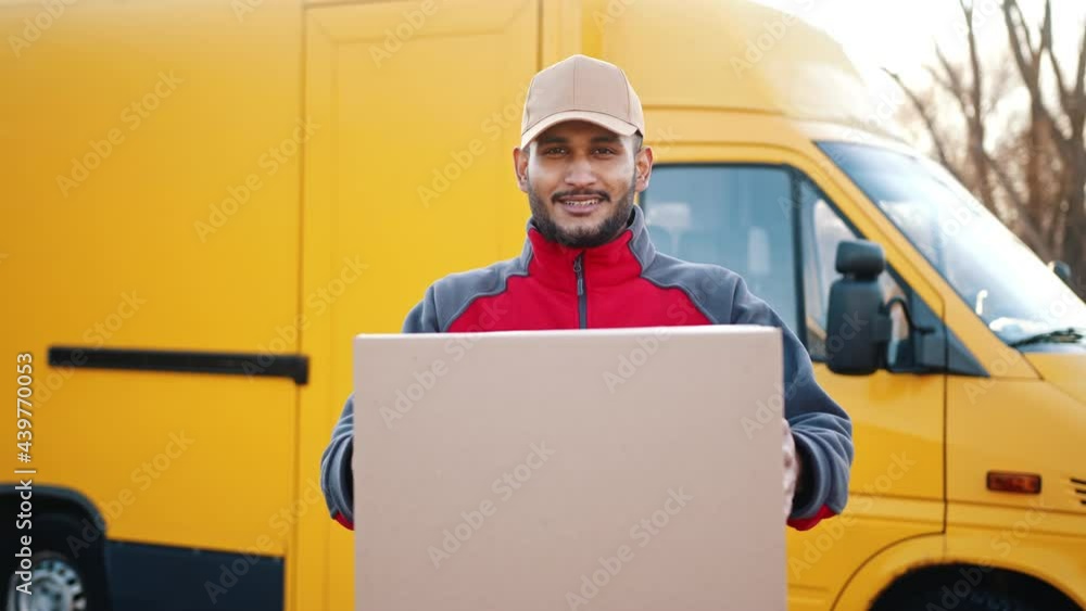 Delivery boy holding a cardboard box in his hands. Smiling and moving the box towards the camera. A yellow truck can be seen in the background. Concept of parcel delivery service. Delivery boy holding a cardboard box in his hands. Smiling and moving the box towards the camera. A yellow truck can be seen in the background. Concept of parcel delivery service.