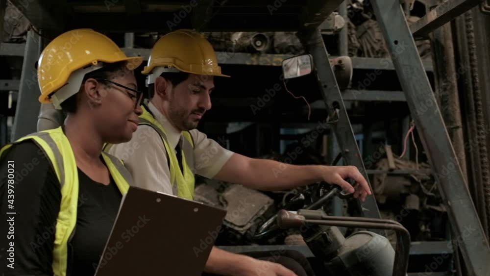 Caucasian male and black female engineers in safety uniforms and helmets on the forklift. Two employees taking about stock order of auto spare parts, engines, and machines in the factory warehouse. Caucasian male and black female engineers in safety uniforms and helmets on the forklift. Two employees taking about stock order of auto spare parts, engines, and machines in the factory warehouse.