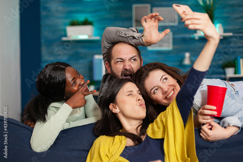 Multi-ethnic friends bonding together taking pictures making diverse expression posting on social media. Group of mixed race people laughting while sitting on sofa late at night in living room.