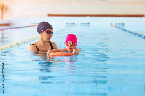 Portrait of mother or coach learning to swim with flutter board little girl at the public swimming-pool. Space for text