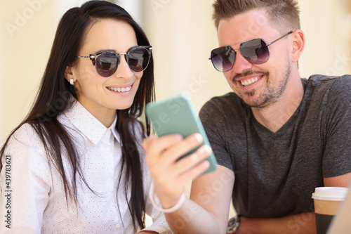 Portrait of man and woman in sunglasses sitting in cafe and looking into smartphone