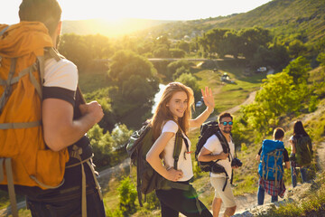 Group of happy backpackers trekking on sunny day. Young tourists traveling and enjoying active summer vacation. Smiling woman looking at camera and waving hand walking down hiking trail with friends