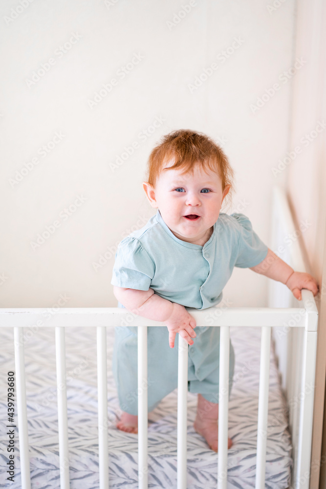 A toddler with red hair is holding onto the side of the crib. Danger for the child to fall out of bed. The baby learns to stand at the support. A toddler with red hair is holding onto the side of the crib. Danger for the child to fall out of bed. The baby learns to stand at the support.