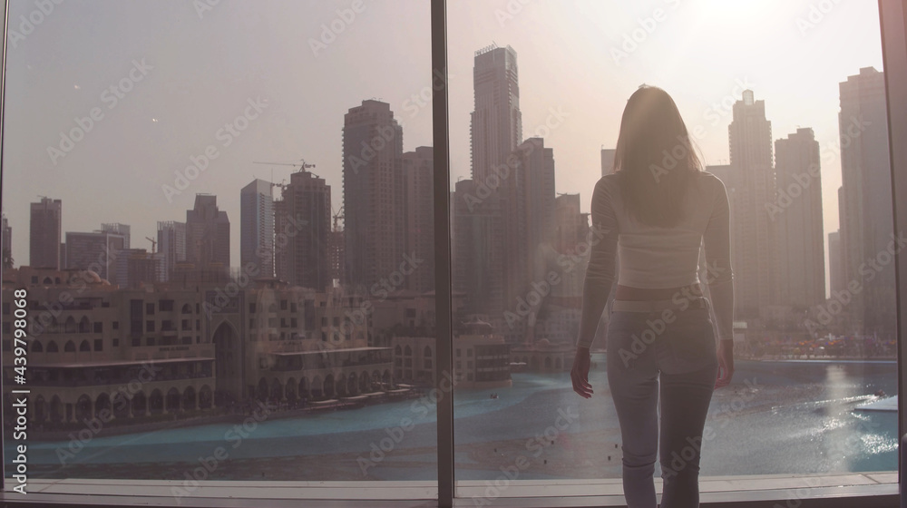 Young woman taking photo at the big window and looking on the Dubai city buildings. Sunset.