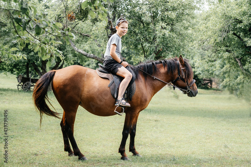 A happy young boy riding a brown horse in the countryside, a horse ranch in the summer © Marinesea