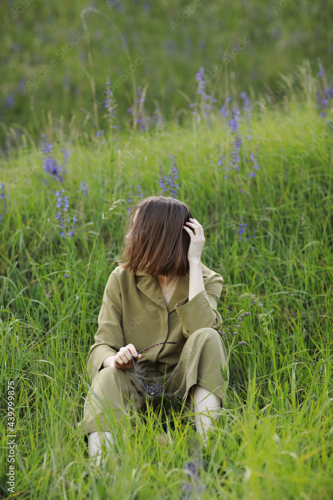 Fototapeta premium Portrait of a young woman sitting in a field on the spring grass among purple flowers. stylish girl enjoys Sunny spring weather. Natural beauty of a woman, natural cosmetics