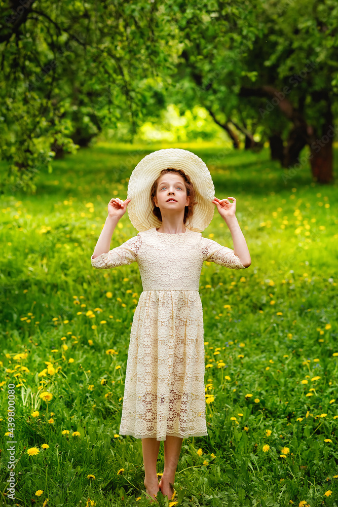 A girl dances among the blooming dandelions and grass in the park. A girl in a hat and a light lace dress in an apple orchard.