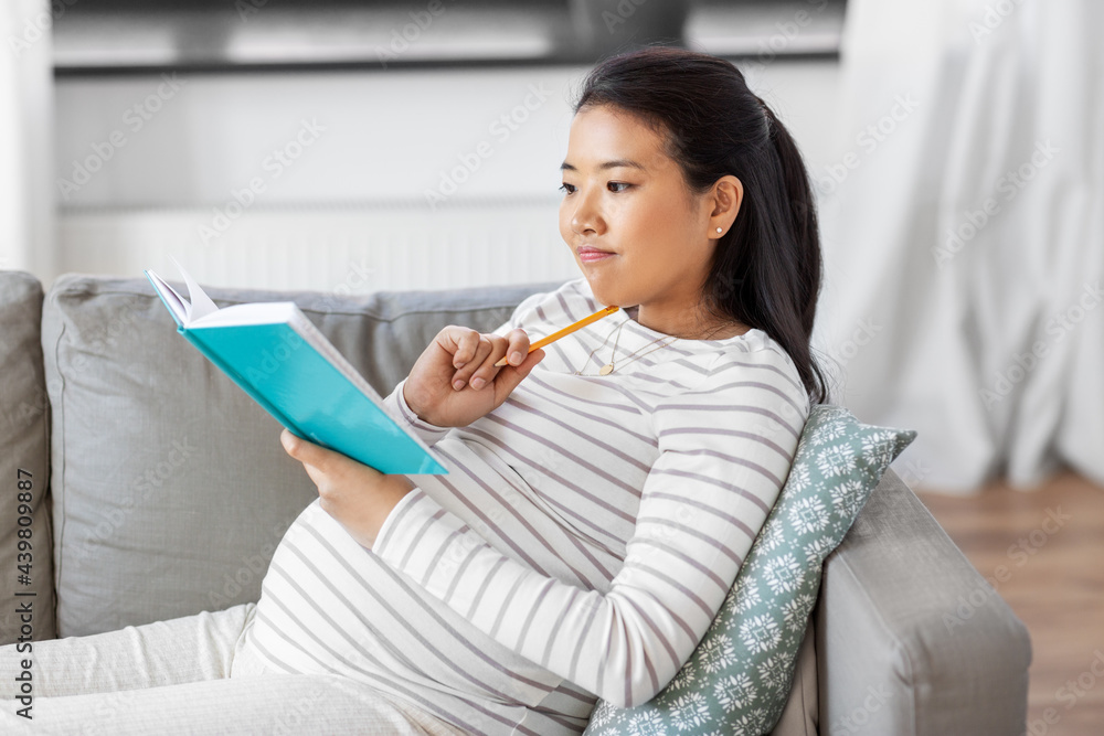 pregnancy, people and leisure concept - pregnant asian woman sitting on sofa and writing to diary or notebook at home pregnancy, people and leisure concept - pregnant asian woman sitting on sofa and writing to diary or notebook at home