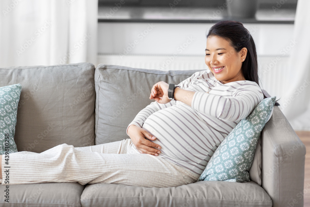 pregnancy, rest, people and expectation concept - happy smiling pregnant asian woman with smart watch sitting on sofa at home pregnancy, rest, people and expectation concept - happy smiling pregnant asian woman with smart watch sitting on sofa at home