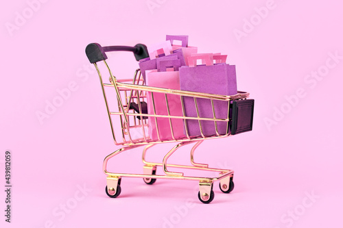 Shopping cart filled with pink and purple paper shopping bags on pink background