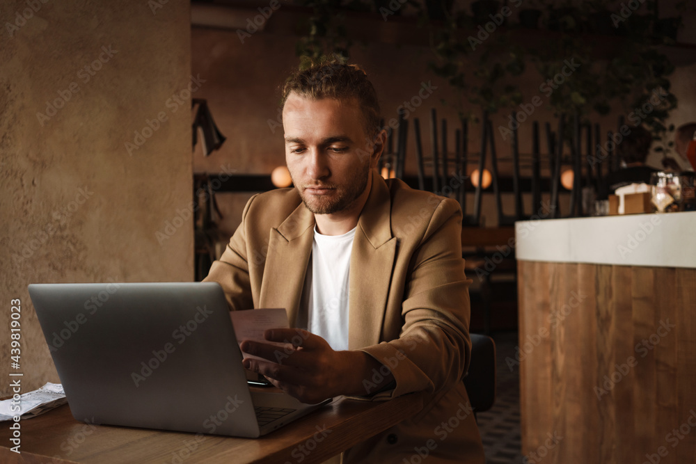 Young white man working with laptop and receipt while sitting in cafe Young white man working with laptop and receipt while sitting in cafe