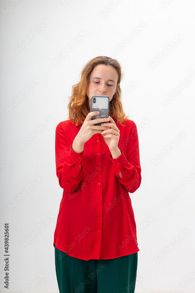young concentrated caucasian redheaded pretty woman with long hair in red shirt isolated on white background holds smartphone in hands, takes picture. Technology, communication, using mobile phone young concentrated caucasian redheaded pretty woman with long hair in red shirt isolated on white background holds smartphone in hands, takes picture. Technology, communication, using mobile phone