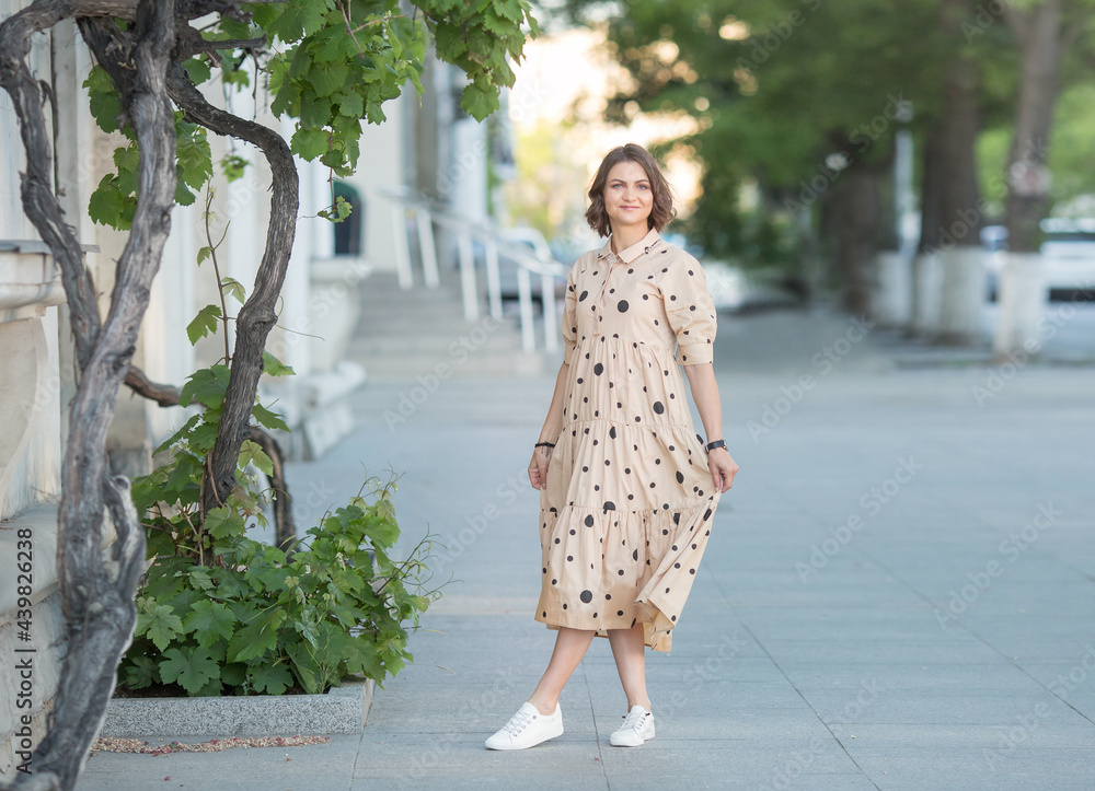 a happy girl stands in a dress on a green street of the city and smiles a happy girl stands in a dress on a green street of the city and smiles