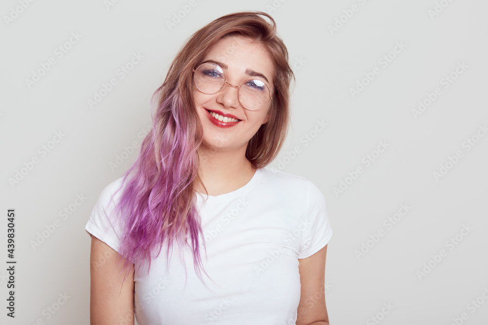 Pleasant looking female in glasses and white casual T-shirt looking smiling directly at camera, has lilac hair, expressing positive emotions, isolated over grey background. Pleasant looking female in glasses and white casual T-shirt looking smiling directly at camera, has lilac hair, expressing positive emotions, isolated over grey background.