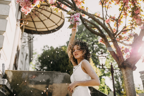 Young woman with curly hair, posing outdoors, next to a tree in bloom with pink flowers and petals falling.