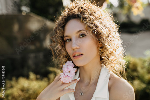 Portrait of a young woman with curly hair, posing outdoors with pink flowers.