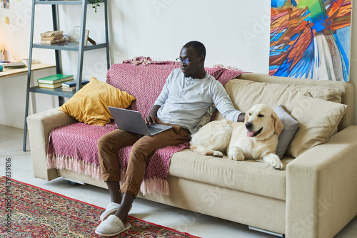 African man sitting on sofa with his dog and using laptop for online work in domestic room