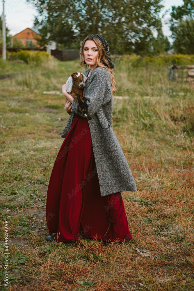 A beautiful woman in a long coat and a red dress holds a little goat in her arms.