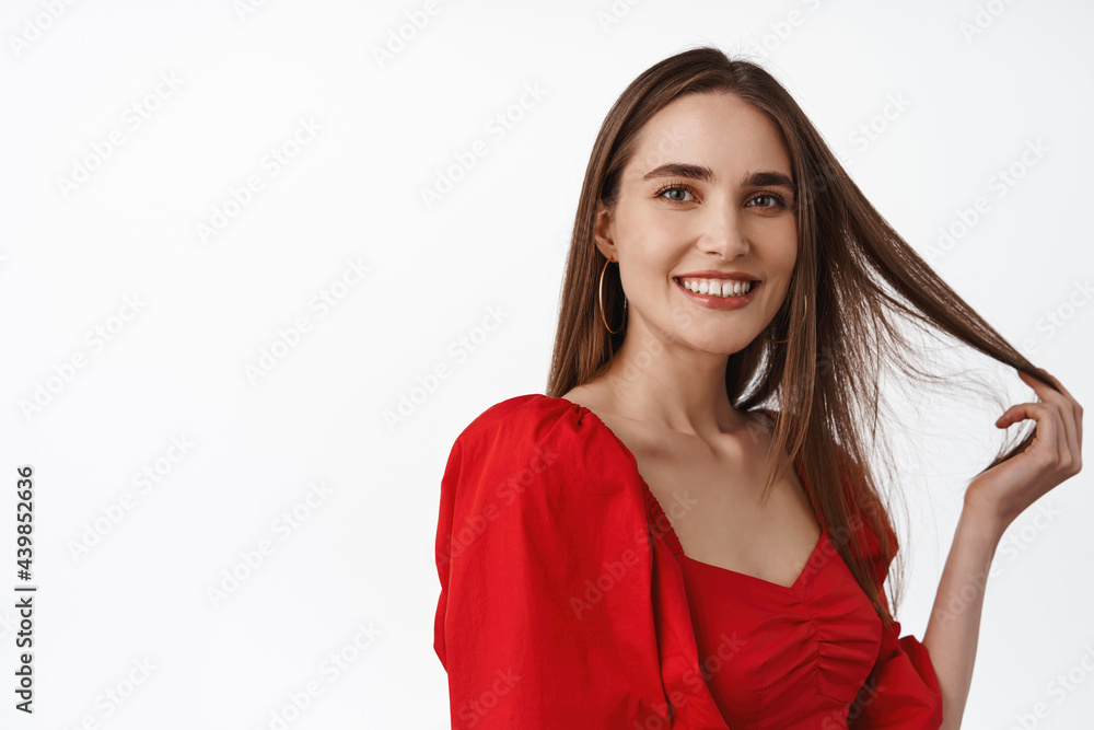 Close up portrait of attractive and confident woman playing with hair strand, look flirty and smiling at camera, standing in red dress against white background Close up portrait of attractive and confident woman playing with hair strand, look flirty and smiling at camera, standing in red dress against white background