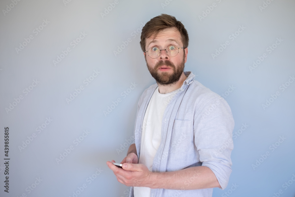 Confused, shocked man with a smartphone in hands looks at the camera in surprise, with big eyes. Gray background. Confused, shocked man with a smartphone in hands looks at the camera in surprise, with big eyes. Gray background.