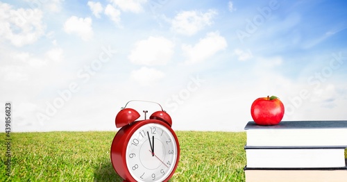 Apple over stack of books and red alarm clock against landscape with green grass and blue sky