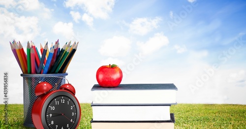 Apple over stack of books, alarm clock and pencil stand against landscape with grass and blue sky