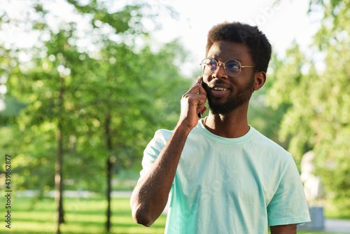 African young man in eyeglasses talking on mobile phone and smiling while standing in the park © AnnaStills