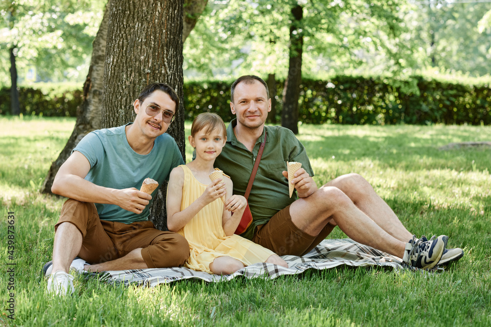 Portrait of gay couple sitting on the grass with child and eating ice creams together Portrait of gay couple sitting on the grass with child and eating ice creams together