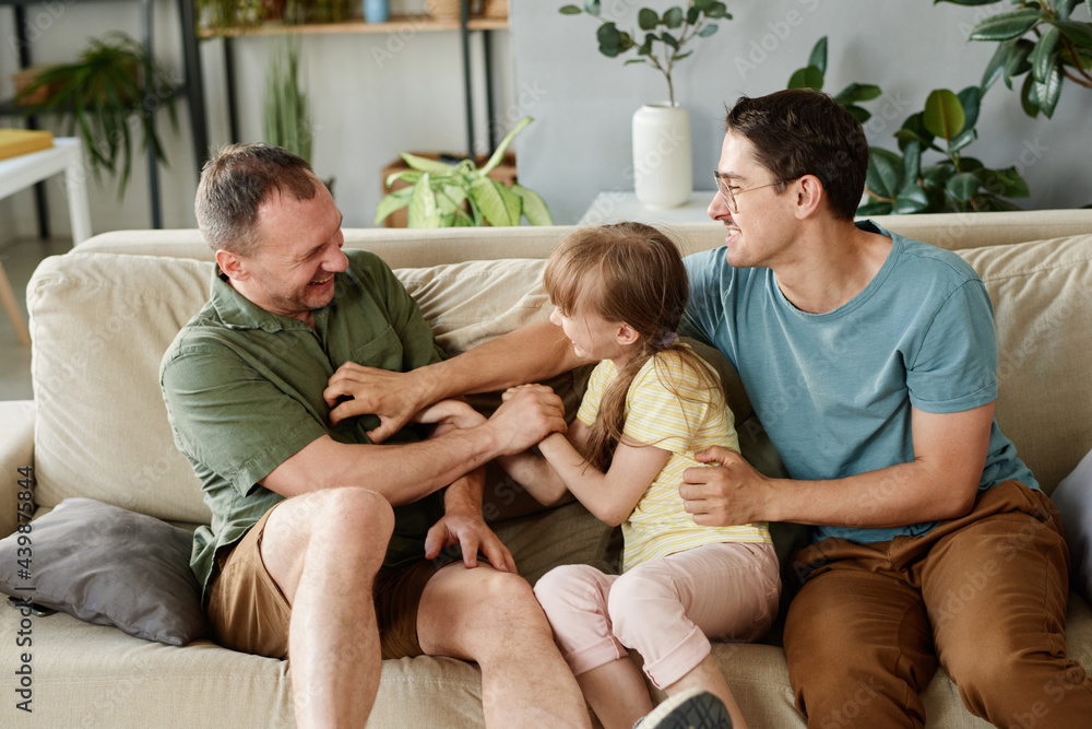 Happy gay family playing together with adopted daughter on the sofa at home
