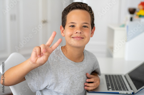Smiling young boy in bedroom using laptop with victory sign gesture