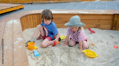 Little boy digs sand with green rake spending time with sister on contemporary playground in park