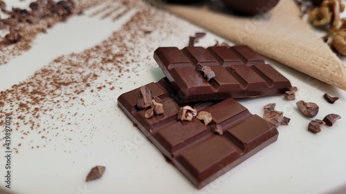 Two milk chocolate bars in white plate, in composition with raw cocoa powder and fork print, candies with chocolate and chocolate drops and shavings. Walnuts and tissue for decoration. 