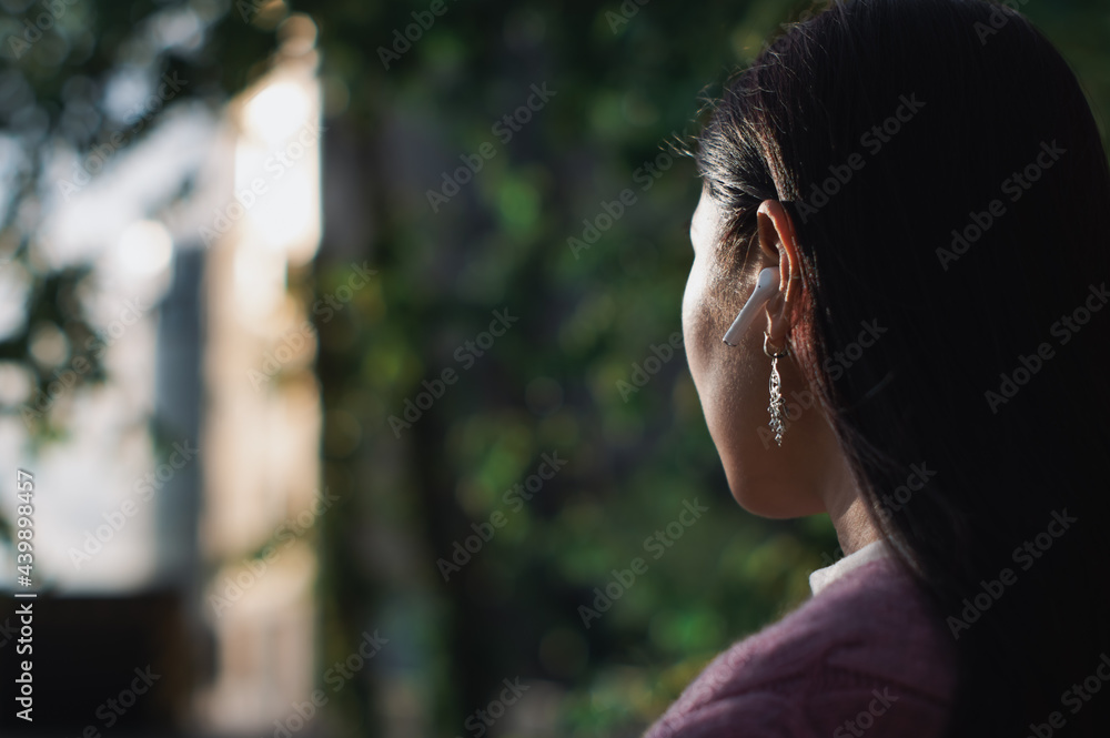 rear view of asian woman in casual clothes in city with wireless earphone in ear looking at sun. close-up against blurred background
