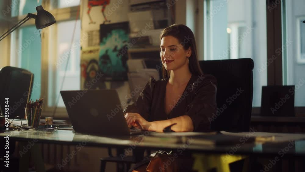 Portrait of a Smiling Young Beautiful Female Working on a Laptop Computer in Creative Agency in Loft Office. Renovated Stylish Design with House Plants, Artistic Posters and Big Rounded Windows. Portrait of a Smiling Young Beautiful Female Working on a Laptop Computer in Creative Agency in Loft Office. Renovated Stylish Design with House Plants, Artistic Posters and Big Rounded Windows.