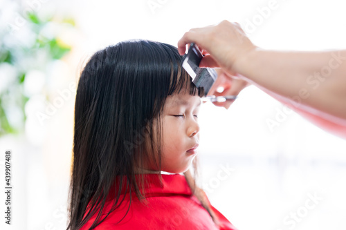 Cute Asian girl sitting and eyes closed. The barber's hands skillfully cut front hair of child. Kid cover their bodies with red veils to prevent hair from sticking on clothes. Children are 4 years old