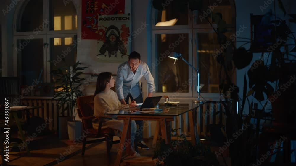 Art Director and Marketing Manager Discussing Work-Related Projects on a Laptop Computer in Creative Agency in Loft Office. Dark Renovated Space in the Evening with Plants, Posters and Big Windows. Art Director and Marketing Manager Discussing Work-Related Projects on a Laptop Computer in Creative Agency in Loft Office. Dark Renovated Space in the Evening with Plants, Posters and Big Windows.