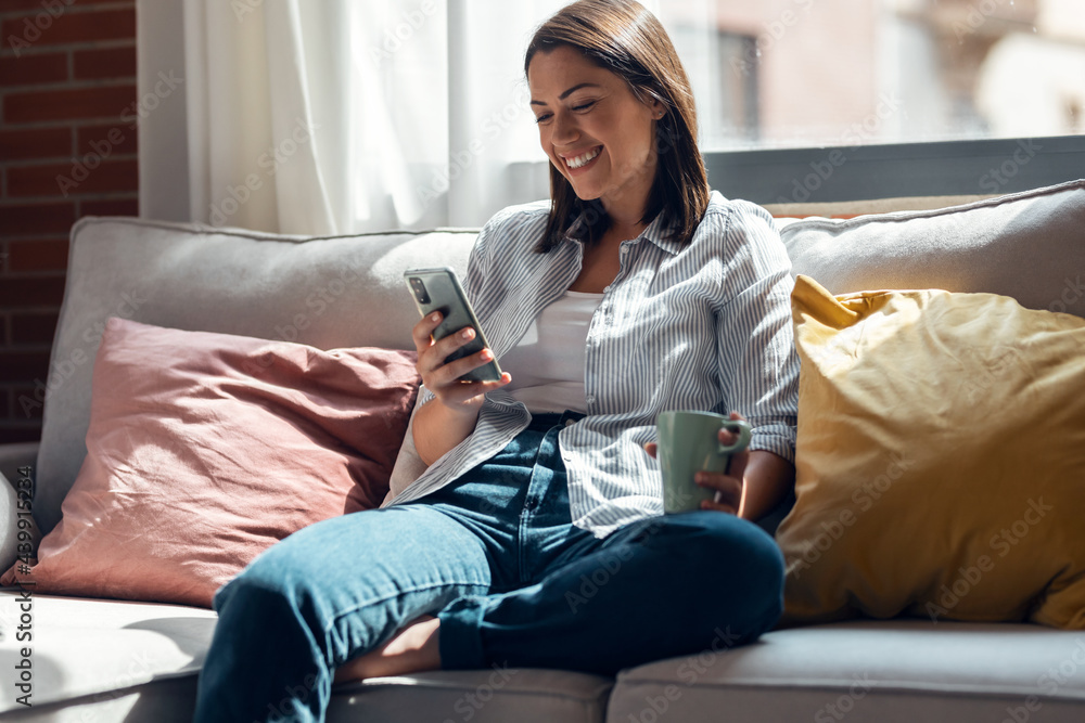 Pretty young smiling woman using mobile phone sitting on a couch at home. - obrazy, fototapety, plakaty 
