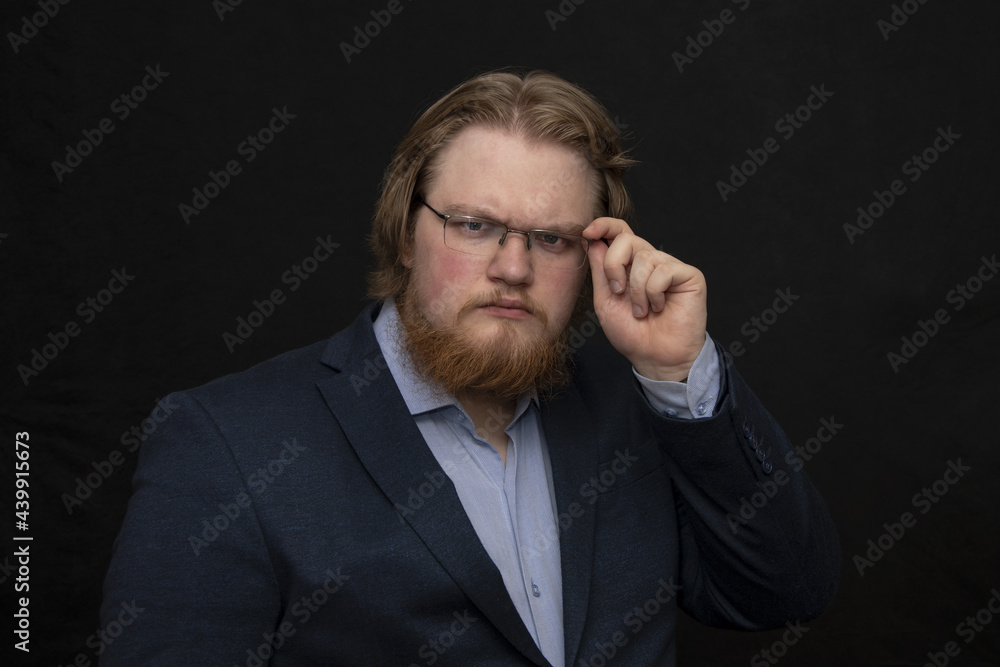 Portrait of a serious man 25-30 years old with glasses and a suit, with a red beard on a dark background looking at the camera, overweight, fat,