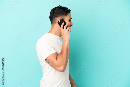 Young arab man isolated on blue background keeping a conversation with the mobile phone with someone