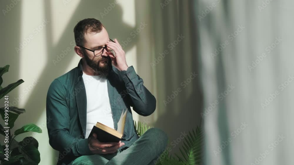 Bearded young man sitting near window and reading a book at the sunrise, enjoy the moment and admire the view. Sun illuminate his face.