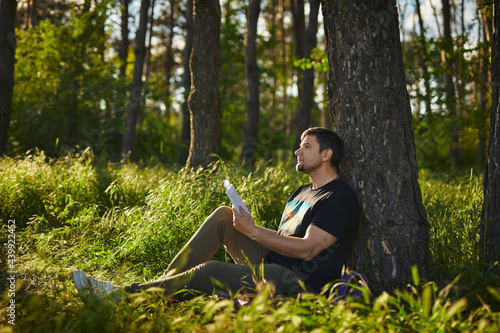 Handsome young man sitting in the forest on the grass reading a book