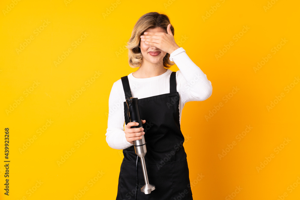 Girl using hand blender isolated on yellow background covering eyes by hands and smiling