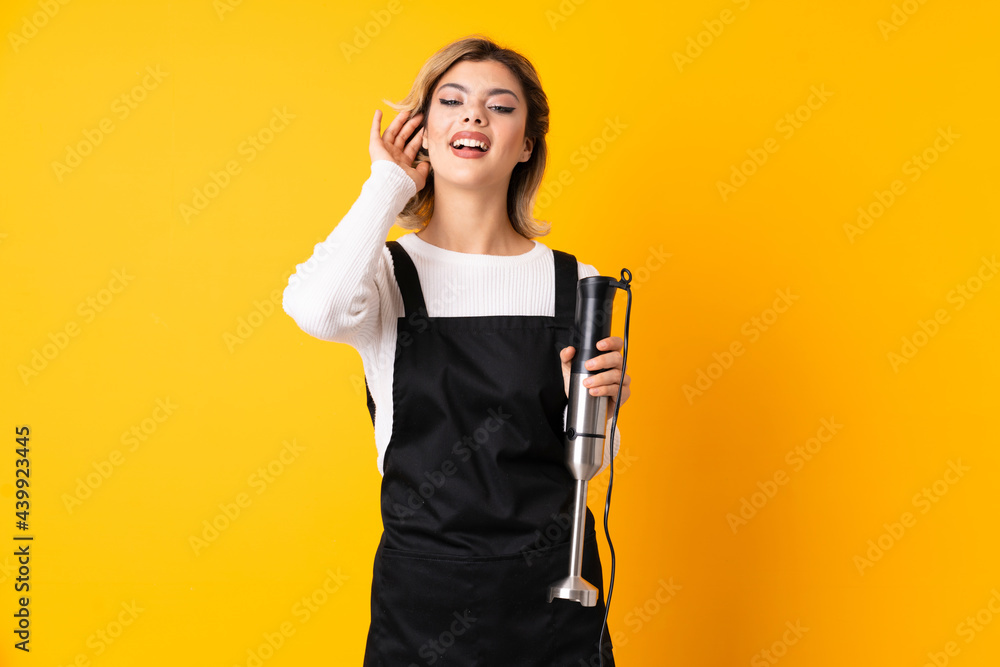 Girl using hand blender isolated on yellow background listening to something by putting hand on the ear Girl using hand blender isolated on yellow background listening to something by putting hand on the ear