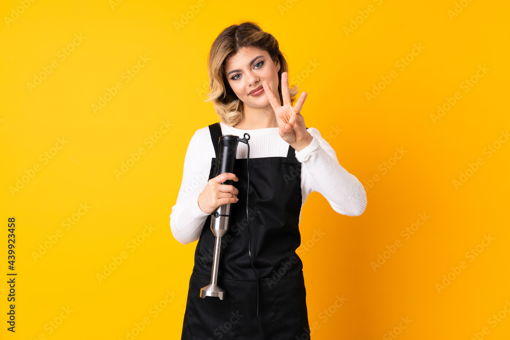 Girl using hand blender isolated on yellow background happy and counting three with fingers Girl using hand blender isolated on yellow background happy and counting three with fingers