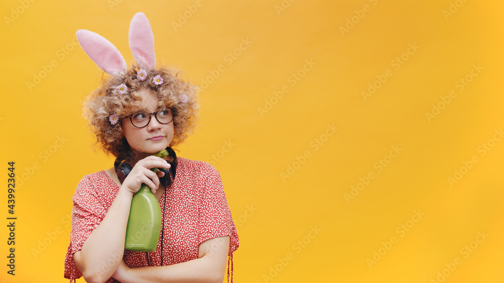 Joyful girl wearing cute bunny ears hairband and spectacles. Holding a green spray bottle. Headphones around her neck. Isolated on yellow background. Easter concept. Joyful girl wearing cute bunny ears hairband and spectacles. Holding a green spray bottle. Headphones around her neck. Isolated on yellow background. Easter concept.
