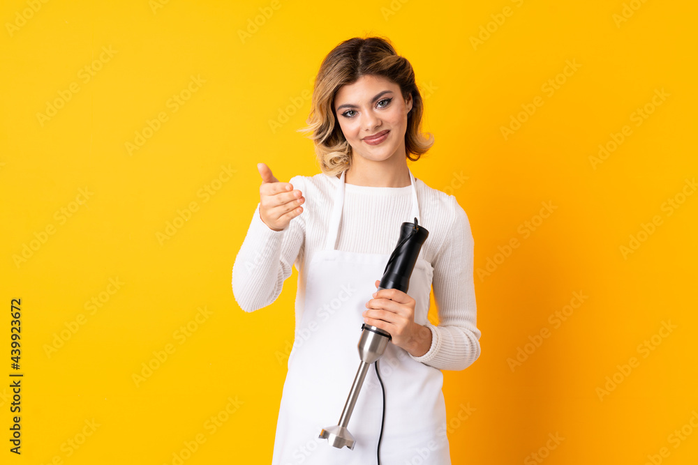 Girl using hand blender isolated on yellow background shaking hands for closing a good deal Girl using hand blender isolated on yellow background shaking hands for closing a good deal