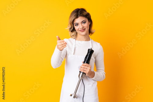 Girl using hand blender isolated on yellow background shaking hands for closing a good deal