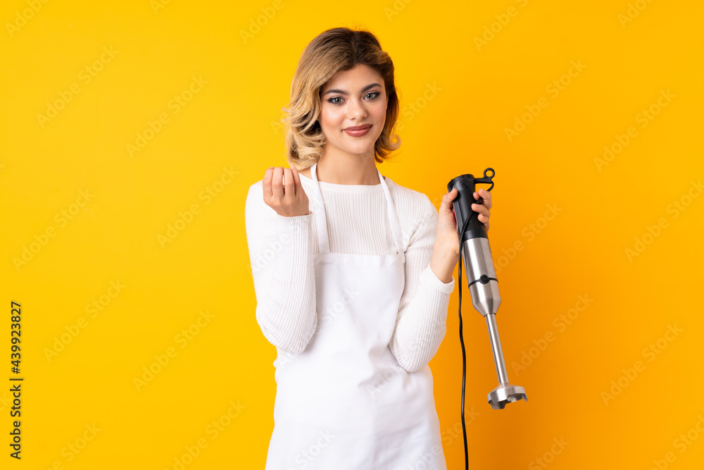 Girl using hand blender isolated on yellow background making money gesture Girl using hand blender isolated on yellow background making money gesture