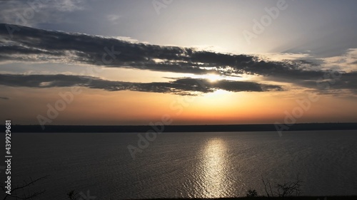 Golden sunset at river shore with sun reflection in rippled water. Sun rays break through dark grey clouds in sky. Tranquil landscape with nobody photo